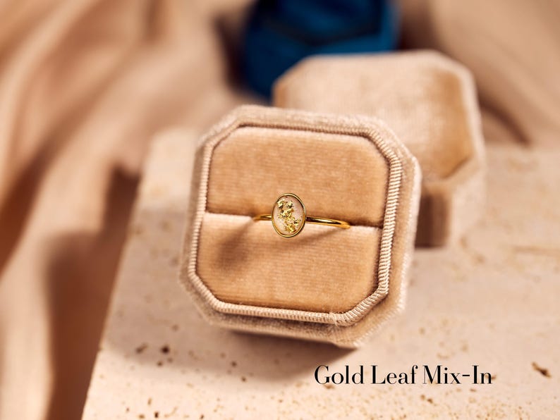 May include: A gold ring with an oval-shaped centerpiece containing gold leaf details, presented in a beige velvet ring box. The ring has a delicate band and is set against a neutral background. The text "Gold Leaf Mix-In" is visible.
