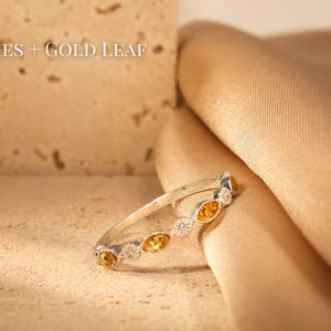 May include: A silver ring with alternating yellow and clear gemstones. The ring is set against a textured beige surface and a draped gold fabric. The text "ASHES + GOLD LEAF" is visible in the upper left corner.