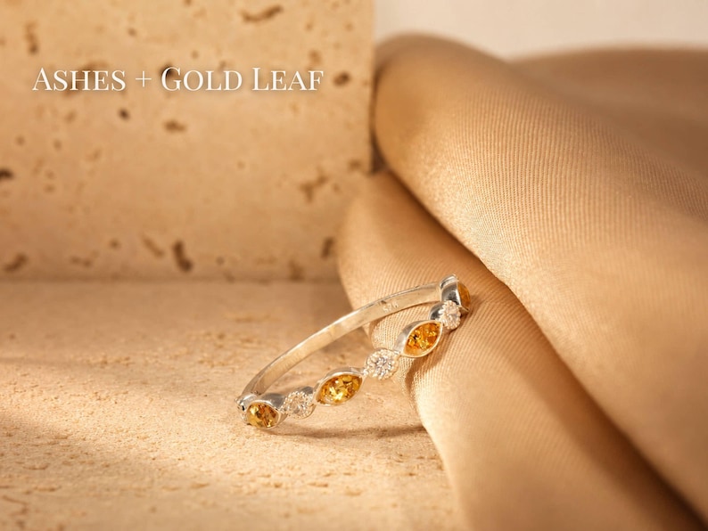 May include: A silver ring with alternating yellow and clear gemstones. The ring is set against a textured beige surface and a draped gold fabric. The text "ASHES + GOLD LEAF" is visible in the upper left corner.