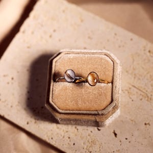 May include: Two rings with round, light-colored stones are displayed in a velvet ring box. One ring has a silver band, and the other has a gold band. The box is beige, and the background is a textured, light-colored surface.