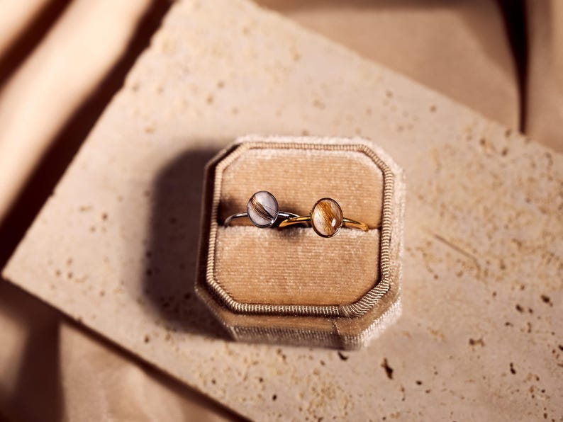 May include: Two rings with round, light-colored stones are displayed in a velvet ring box. One ring has a silver band, and the other has a gold band. The box is beige, and the background is a textured, light-colored surface.