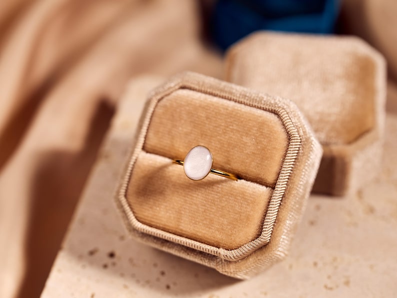 May include: A gold ring with an oval, light-colored stone set in a velvet ring box. The ring is displayed in a beige velvet box, with another box in the background. The background has a soft, neutral color palette.