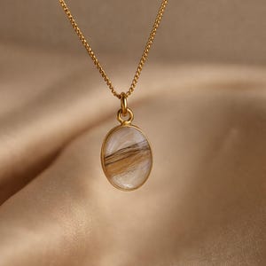 May include: A gold-colored necklace with a pendant. The pendant is oval-shaped, with a gold border, and contains a mix of white and brown hair. The chain is delicate and also gold-colored. The background is a soft, neutral fabric.