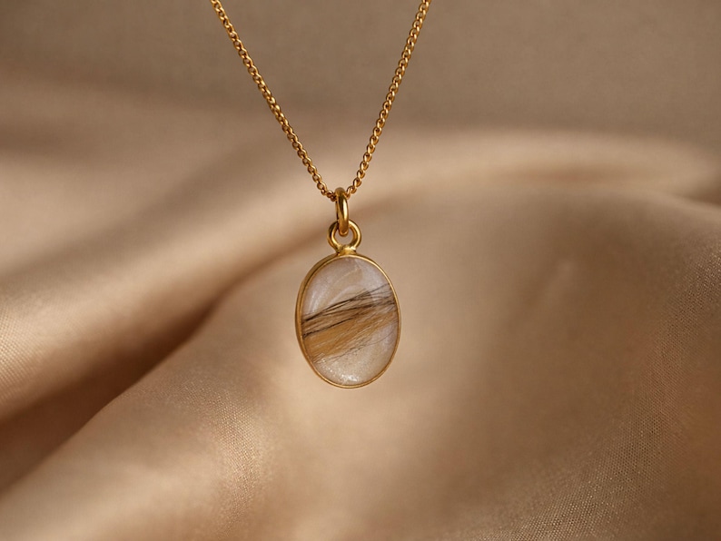 May include: A gold-colored necklace with a pendant. The pendant is oval-shaped, with a gold border, and contains a mix of white and brown hair. The chain is delicate and also gold-colored. The background is a soft, neutral fabric.