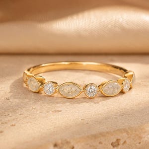 May include: A gold ring with alternating oval and round gemstones. The ring features a series of oval-shaped, light-colored stones interspersed with round, clear gemstones. The band is a polished gold color, and the ring is set against a neutral background.