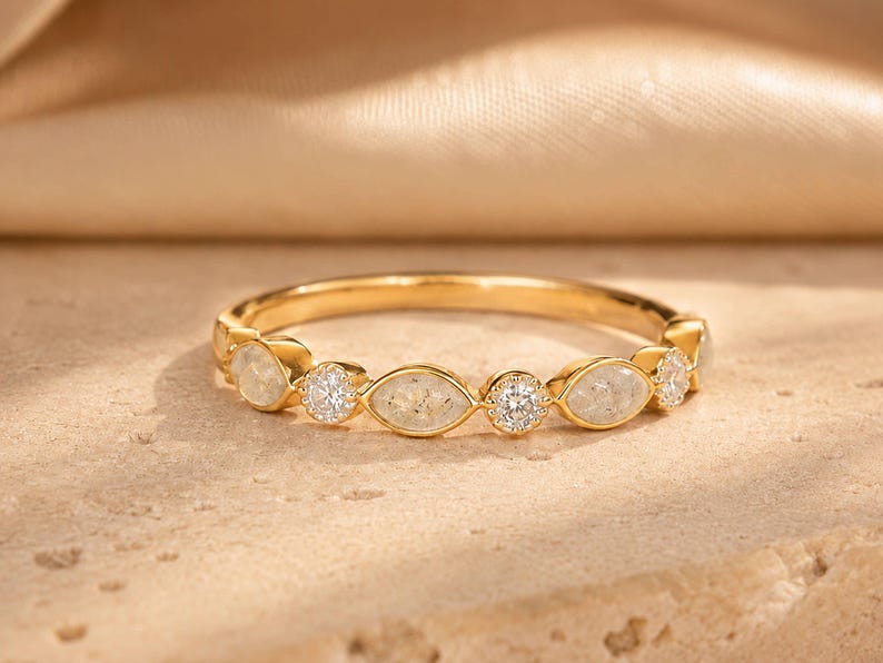 May include: A gold ring with alternating oval and round gemstones. The ring features a series of oval-shaped, light-colored stones interspersed with round, clear gemstones. The band is a polished gold color, and the ring is set against a neutral background.