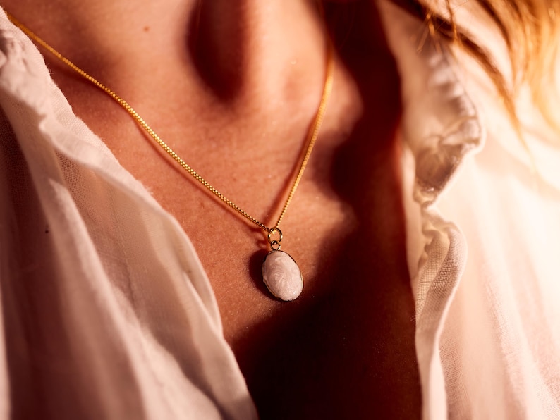 May include: A gold-colored chain necklace with a small, oval, white pendant. The necklace is worn against a white shirt. The pendant has a textured surface and is the focal point of the image.