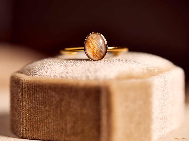 May include: A gold ring with an oval-shaped, amber-colored stone. The ring is set on a beige velvet ring box. The stone has a unique, natural pattern with golden and brown inclusions. The ring's band is thin and delicate.
