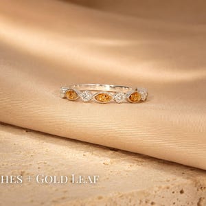 May include: A silver ring with alternating orange and clear gemstones. The ring is set on a beige fabric surface. The text "Ashes + Gold Leaf" is at the bottom of the image.