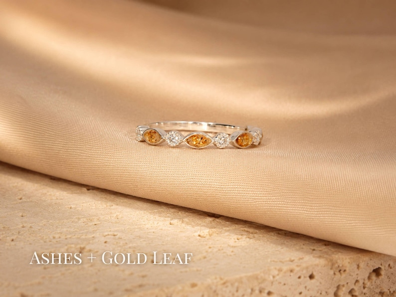 May include: A silver ring with alternating orange and clear gemstones. The ring is set on a beige fabric surface. The text "Ashes + Gold Leaf" is at the bottom of the image.