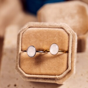 May include: Two rings with oval, light-colored stones are displayed in a velvet ring box. One ring has a silver band, and the other has a gold band. The background is a soft, neutral color.