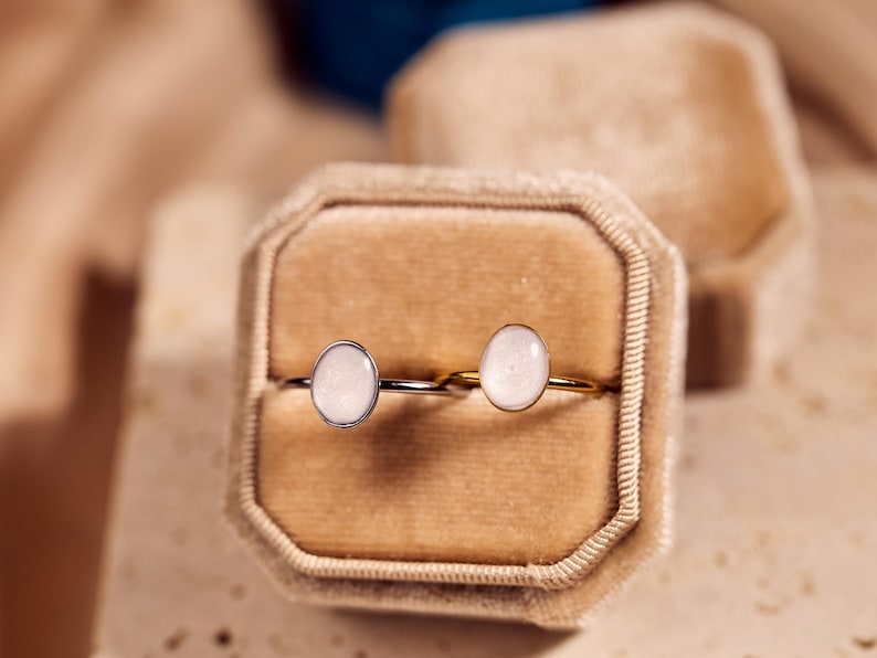 May include: Two rings with oval, light-colored stones are displayed in a velvet ring box. One ring has a silver band, and the other has a gold band. The background is a soft, neutral color.