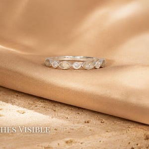 May include: A silver ring with a series of marquise-shaped stones and a central round stone. The ring is set against a tan fabric background. The text "Ashes Visible" is at the bottom of the image.
