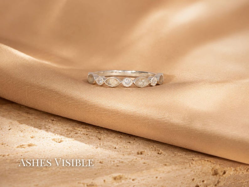 May include: A silver ring with a series of marquise-shaped stones and a central round stone. The ring is set against a tan fabric background. The text "Ashes Visible" is at the bottom of the image.