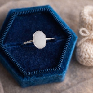 May include: A silver ring with an oval, light-colored stone is displayed in a dark blue velvet ring box. A pair of cream-colored, crocheted baby booties with a bow are next to the ring box. The background is a neutral, light brown fabric.