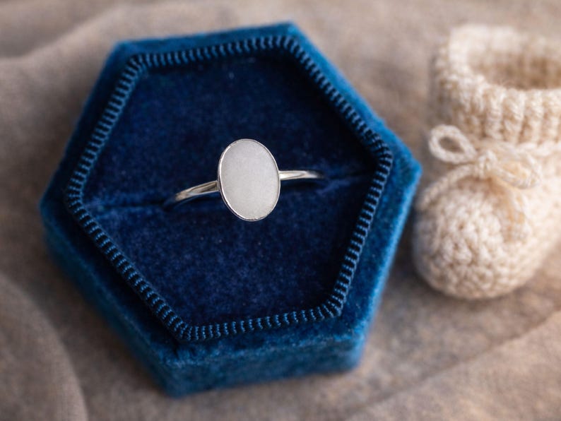 May include: A silver ring with an oval, light-colored stone is displayed in a dark blue velvet ring box. A pair of cream-colored, crocheted baby booties with a bow are next to the ring box. The background is a neutral, light brown fabric.