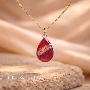 May include: A gold-colored chain necklace with a teardrop-shaped pendant. The pendant is a deep burgundy red with a strand of light-colored hair encased within. The text "Burgundy Red Stone + Hair" is at the bottom of the image.