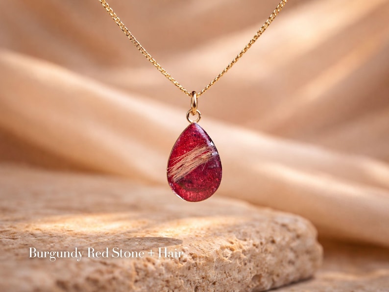 May include: A gold-colored chain necklace with a teardrop-shaped pendant. The pendant is a deep burgundy red with a strand of light-colored hair encased within. The text "Burgundy Red Stone + Hair" is at the bottom of the image.