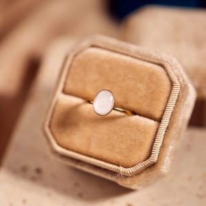 May include: A gold ring with an oval, light-colored stone is displayed in a beige velvet ring box. The ring's band is thin and shiny. The box is open, revealing the ring against a soft, neutral background.