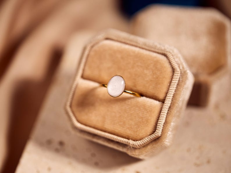 May include: A gold ring with an oval, light-colored stone is displayed in a beige velvet ring box. The ring's band is thin and shiny. The box is open, revealing the ring against a soft, neutral background.