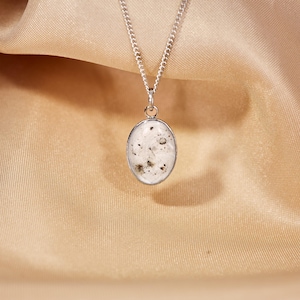 May include: Silver chain necklace with an oval pendant. The pendant has a white background with dark specks and is encased in a silver frame. The necklace is displayed on a gold-colored satin fabric.