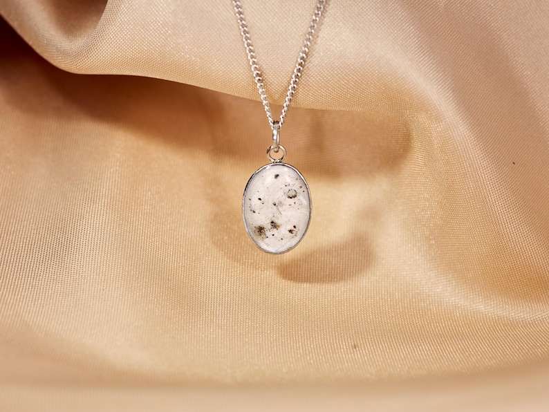 May include: Silver chain necklace with an oval pendant. The pendant has a white background with dark specks and is encased in a silver frame. The necklace is displayed on a gold-colored satin fabric.