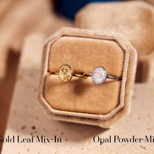 May include: Two rings with oval-shaped stones are displayed in a velvet ring box. One ring has a gold band with a gold leaf mix-in stone, and the other has a silver band with an opal powder mix-in stone. Text at the bottom reads "Gold Leaf Mix-In" and "Opal Powder Mix-In".
