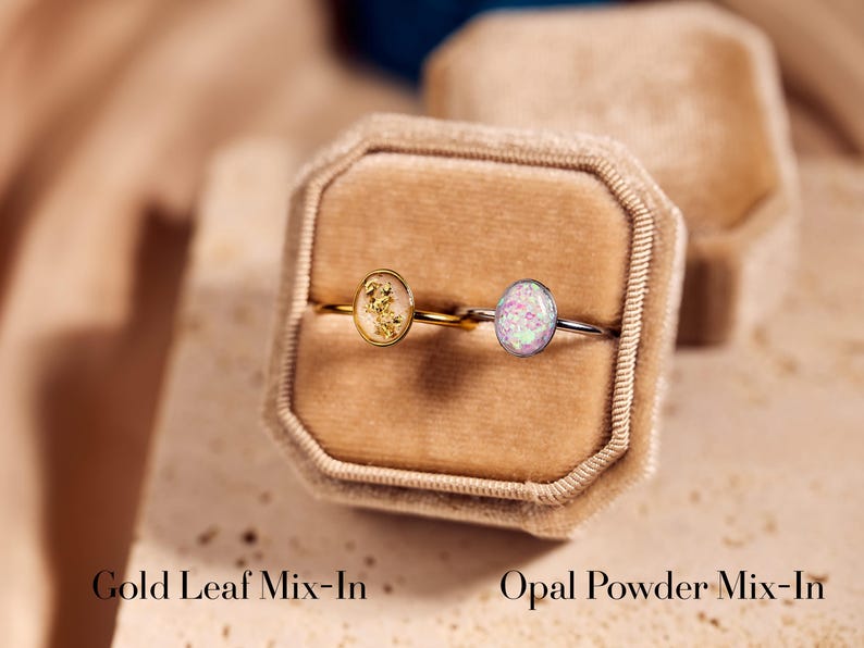 May include: Two rings with oval-shaped stones are displayed in a velvet ring box. One ring has a gold band with a gold leaf mix-in stone, and the other has a silver band with an opal powder mix-in stone. Text at the bottom reads "Gold Leaf Mix-In" and "Opal Powder Mix-In".