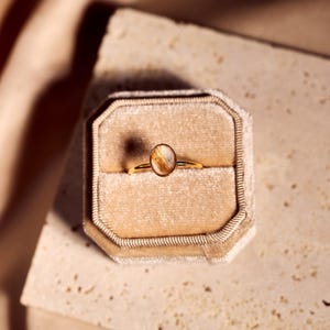 May include: A gold ring with an oval-shaped, light brown gemstone is displayed inside a beige velvet ring box. The ring is set against a neutral background, with soft lighting.