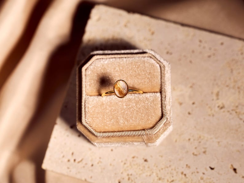 May include: A gold ring with an oval-shaped, light brown gemstone is displayed inside a beige velvet ring box. The ring is set against a neutral background, with soft lighting.