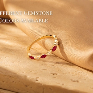 May include: A gold ring with alternating red gemstones and small white stones. The ring is set against a backdrop of a tan fabric and a light-colored surface. Text in the image reads "Different Gemstone Colors Available."
