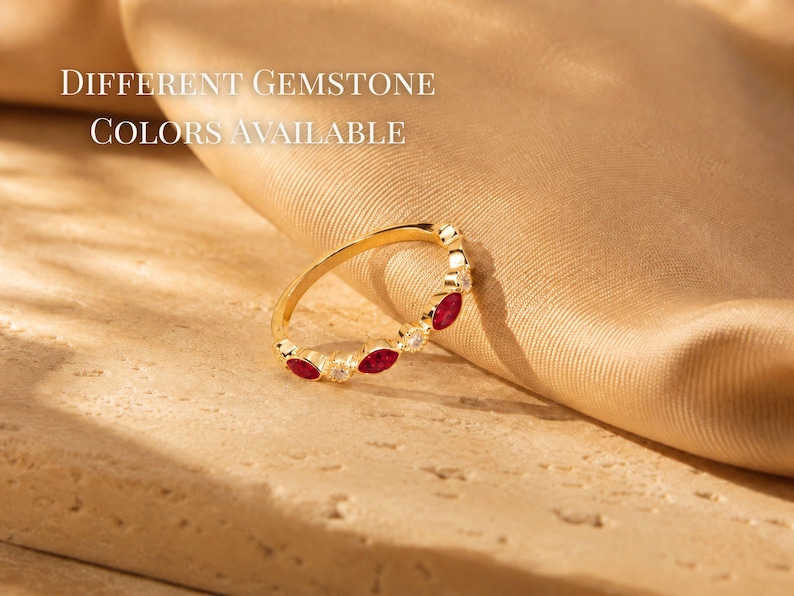 May include: A gold ring with alternating red gemstones and small white stones. The ring is set against a backdrop of a tan fabric and a light-colored surface. Text in the image reads "Different Gemstone Colors Available."