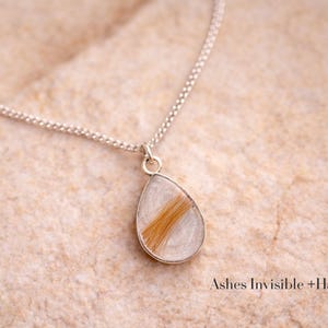 May include: A silver necklace with a teardrop pendant. The pendant contains a section of light brown hair encased in clear resin. The necklace is displayed on a textured, light-colored surface. Text at the bottom reads "Ashes Invisible + Hair."