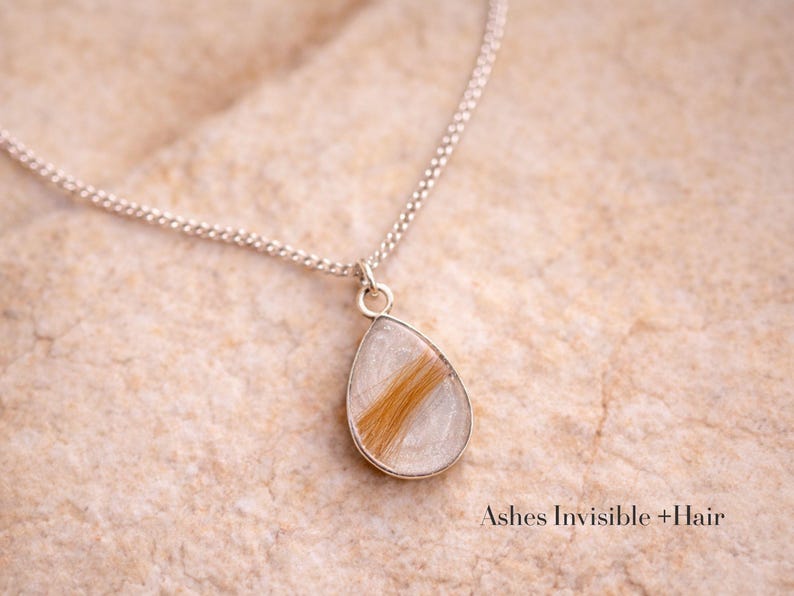May include: A silver necklace with a teardrop pendant. The pendant contains a section of light brown hair encased in clear resin. The necklace is displayed on a textured, light-colored surface. Text at the bottom reads "Ashes Invisible + Hair."