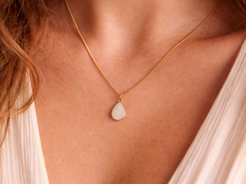 May include: A gold-colored necklace with a teardrop-shaped, off-white pendant. The pendant hangs from a delicate chain, resting against the skin. The necklace is a simple, elegant piece of jewelry.
