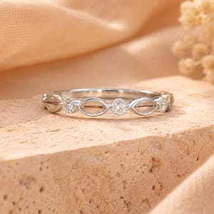 May include: A silver ring with a delicate design. The ring features alternating clear gemstones and open, eye-shaped settings. The gemstones are round and set in a band of silver. The ring is displayed on a textured, light brown surface.