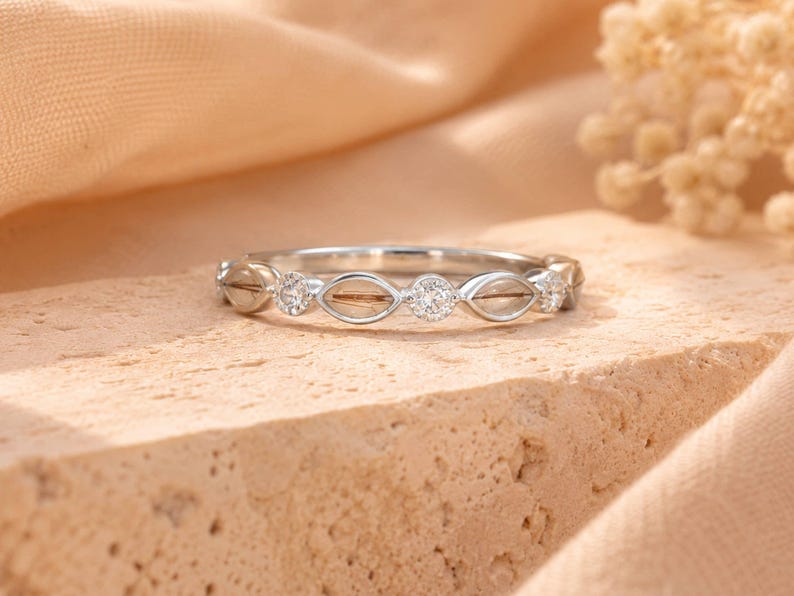 May include: A silver ring with a delicate design. The ring features alternating clear gemstones and open, eye-shaped settings. The gemstones are round and set in a band of silver. The ring is displayed on a textured, light brown surface.