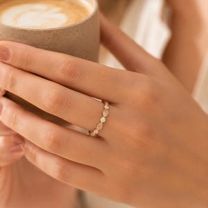 May include: A gold ring with a delicate design featuring alternating marquise and round-cut gemstones. The ring is worn on a finger, and the hand is holding a brown ceramic mug filled with a light-colored beverage.