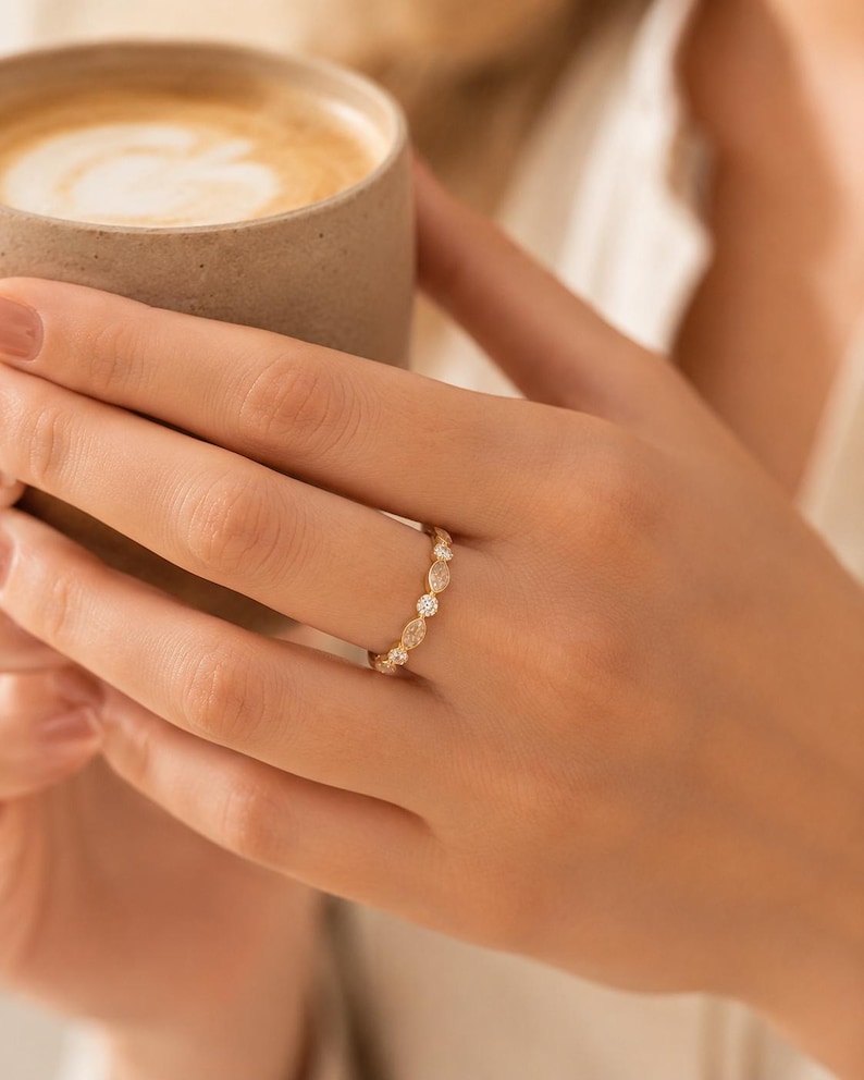 May include: A gold ring with a delicate design featuring alternating marquise and round-cut gemstones. The ring is worn on a finger, and the hand is holding a brown ceramic mug filled with a light-colored beverage.