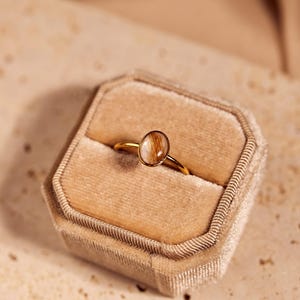 May include: A gold ring with an oval-shaped, light brown stone set in a gold band, displayed in a beige velvet ring box. The ring is centered in the box, showcasing its design and craftsmanship. The background is a neutral beige.