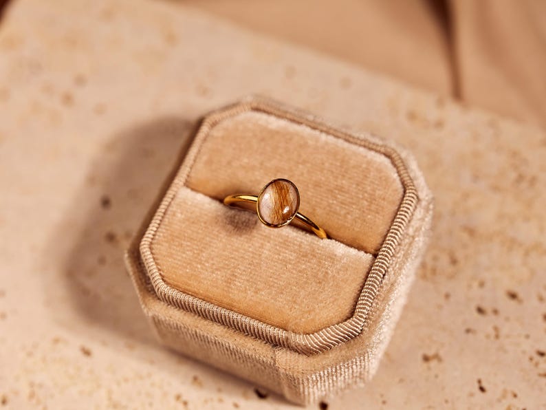 May include: A gold ring with an oval-shaped, light brown stone set in a gold band, displayed in a beige velvet ring box. The ring is centered in the box, showcasing its design and craftsmanship. The background is a neutral beige.
