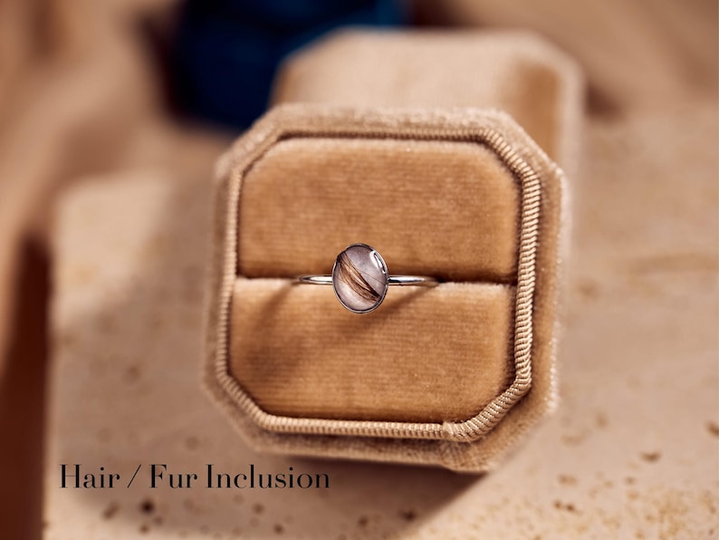 May include: A silver ring with an oval-shaped, clear resin stone containing strands of brown and white hair or fur. The ring is displayed in a velvet ring box. The text "Hair / Fur Inclusion" is at the bottom of the image.