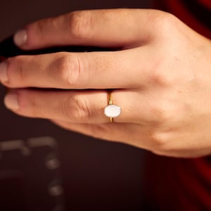 May include: A gold ring with a white oval stone. The ring is on a person's hand, with a dark background and a red and black striped shirt visible in the background.