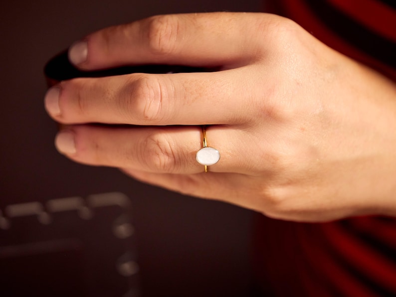 May include: A gold ring with a white oval stone. The ring is on a person's hand, with a dark background and a red and black striped shirt visible in the background.