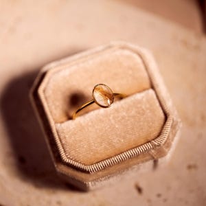 May include: A gold ring with an oval-shaped, clear stone containing strands of brown hair. The ring is displayed in a beige velvet ring box. The ring's band is thin and delicate.