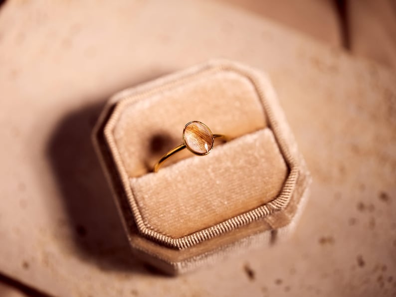 May include: A gold ring with an oval-shaped, clear stone containing strands of brown hair. The ring is displayed in a beige velvet ring box. The ring's band is thin and delicate.