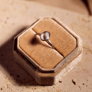 May include: A silver ring with a clear, oval-shaped stone containing brown strands, set in a velvet ring box. The ring is centered in the box, which is a light brown color. The background is a neutral, textured surface.