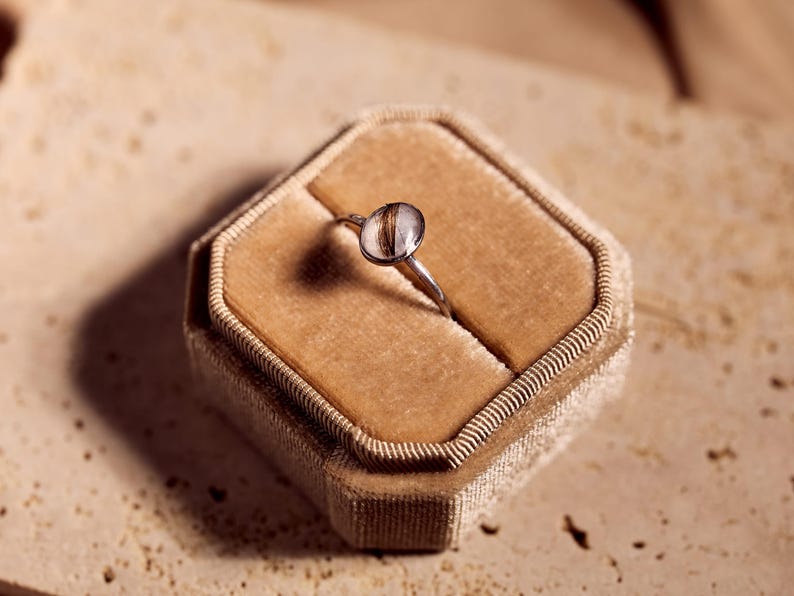 May include: A silver ring with a clear, oval-shaped stone containing brown strands, set in a velvet ring box. The ring is centered in the box, which is a light brown color. The background is a neutral, textured surface.