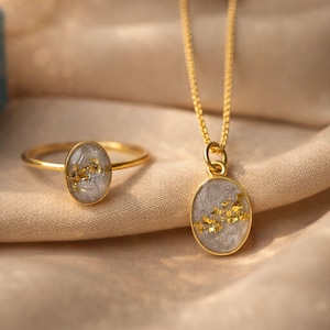 May include: A gold ring and pendant necklace set. The ring and pendant feature an oval-shaped, light gray stone with gold flecks. The jewelry is set against a beige fabric background.