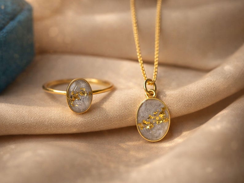 May include: A gold ring and pendant necklace set. The ring and pendant feature an oval-shaped, light gray stone with gold flecks. The jewelry is set against a beige fabric background.